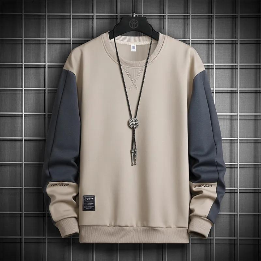 Stijlvolle beige en grijze trui met lange mouwen, ronde hals en contrasterende kleuren. Een ketting met ronde hanger hangt over de trui, wat een elegante touch toevoegt aan het casual kledingstuk.