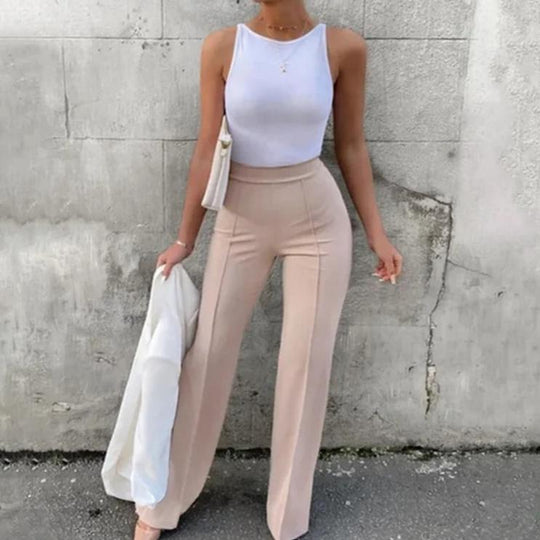 Vrouw draagt elegante flared broek met hoge taille in beige, gecombineerd met een strakke witte crop top. De outfit toont een modieuze en vrouwelijke look, perfect voor diverse gelegenheden. De broek accentueert de silhouet en biedt een comfortabele pasvorm.